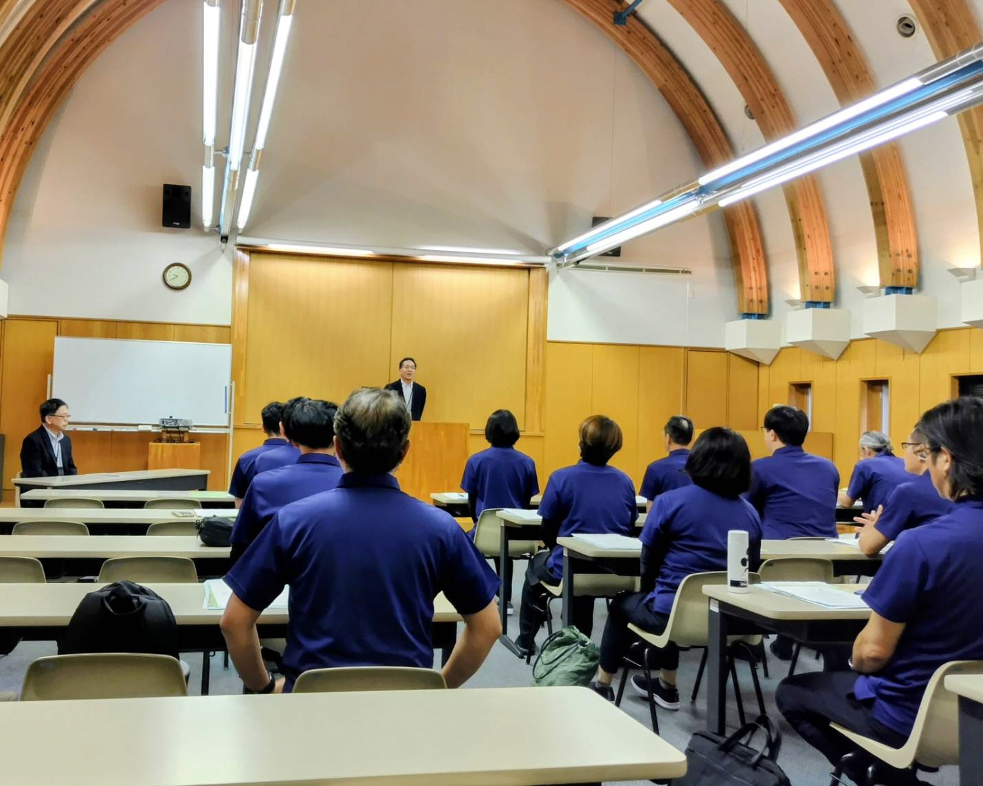 修了式の様子です。農業大学校長の来賓挨拶を行っています。訓練生はお揃いのポロシャツで出席しています。